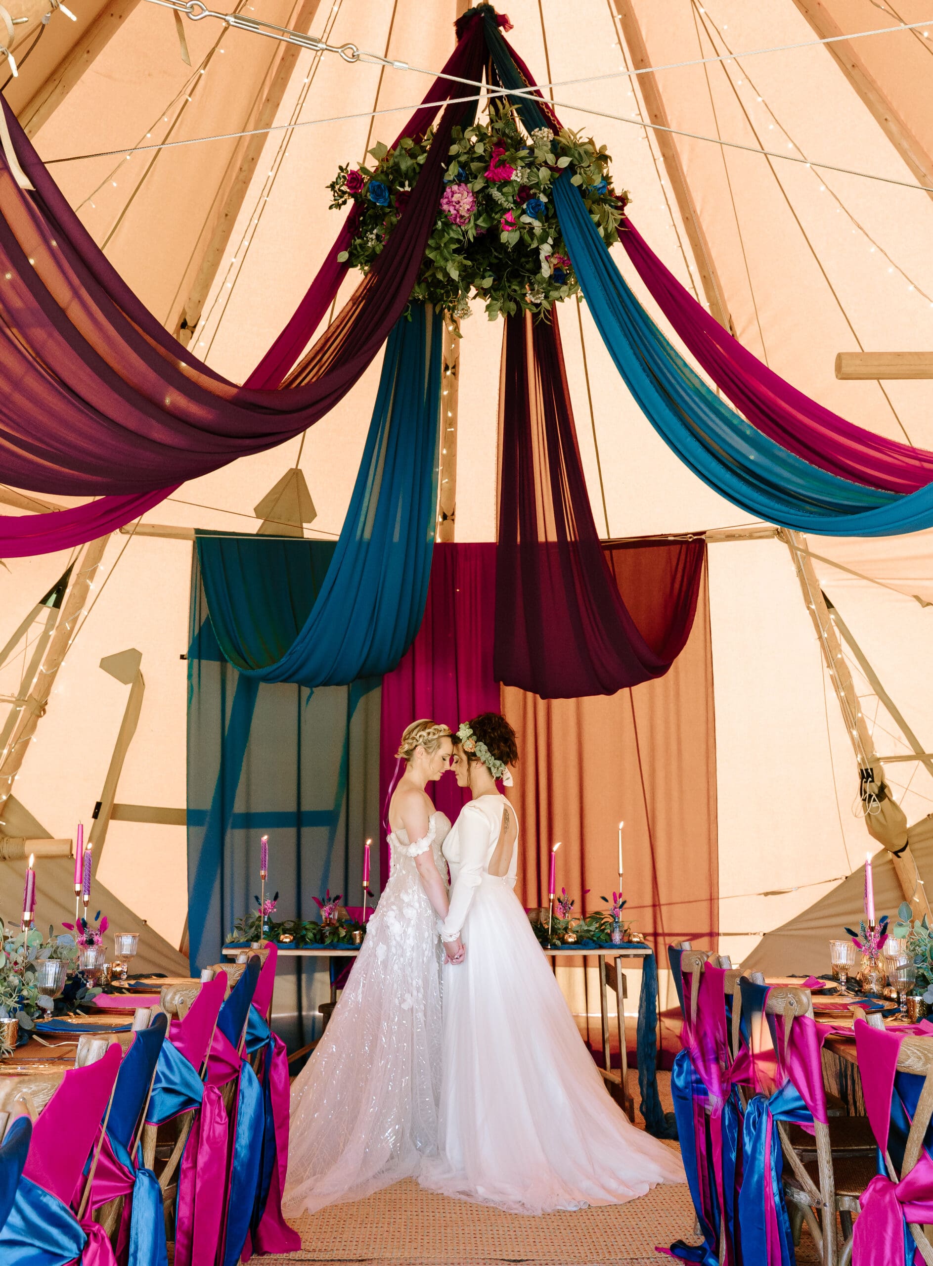 A photo of two brides at their wedding, in a tipi wedding venue under coloured fabric drapes, with styled tables either side.